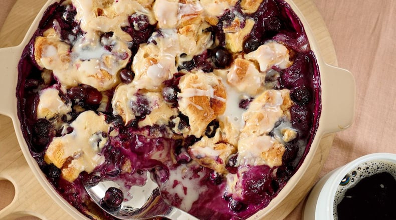 An overhead shot of a blueberry buckle in a beige casserole dish with a serving spoon in it and a cup of coffee next to it.