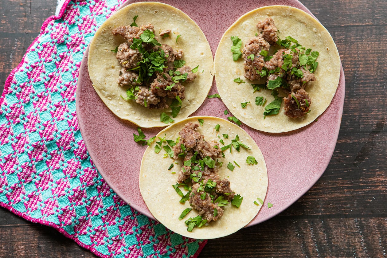 A large serving plate holds three tortillas, each topped with cooked ground pork and garnished with fresh cilantro leaves. The plate is on a wooden table, and a complementary towel is tucked underneath the plate.