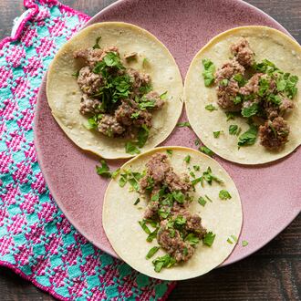 A large serving plate holds three tortillas, each topped with cooked ground pork and garnished with fresh cilantro leaves. The plate is on a wooden table, and a complementary towel is tucked underneath the plate.