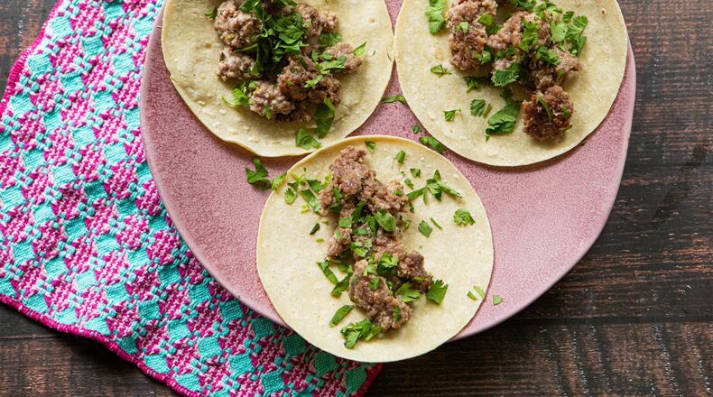 A large serving plate holds three tortillas, each topped with cooked ground pork and garnished with fresh cilantro leaves. The plate is on a wooden table, and a complementary towel is tucked underneath the plate.