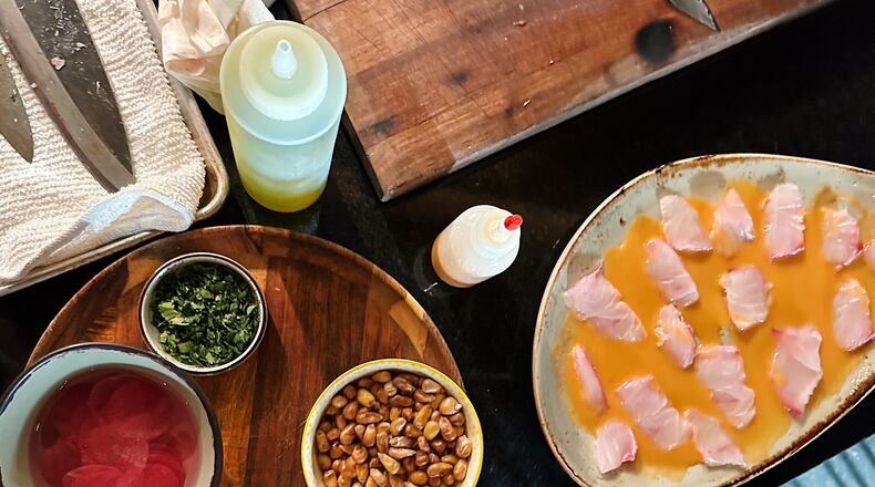 Ingredients for a homemade ceviche are laid out on a table, including a bowl of corn nuts and a plate with fish sliced in thin strips and placed in an orange leche de tigre.