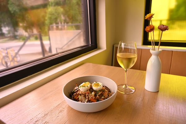 A bowl of fried rice with a soft-boiled egg sitting on top, with a glass of wine and small vase of flowers to the right.