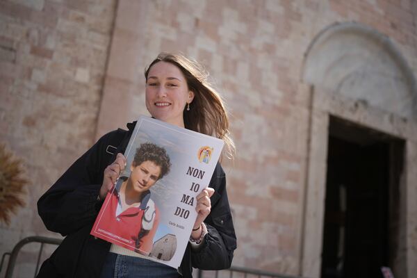Amelia Simone, 18, a student from the United States poses with a banner reading "Not me but God" and a photo of Carlo Acutis, a 15-year-old Italian boy who died in 2006 of leukemia and beatified in 2020, in Assisi, Italy, Wednesday, April 2, 2025. (AP Photo/Alessandra Tarantino)