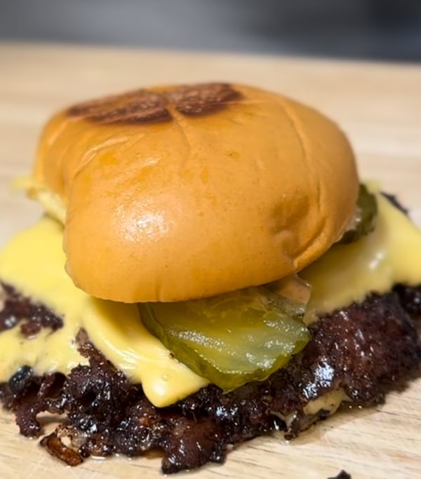 A smashburger made with two patties that hang over the bottom bun, melted American cheese and pickle slices, on a plain bun. The top bun has a brown mark on it like the bun was seared in a skillet or on a blackstone.