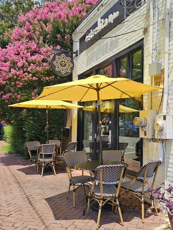 The outside of a restaurant on a sunny day; two tables outside the front door of the brick building are shaded by umbrellas.