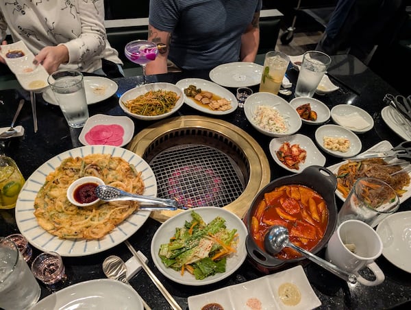 A diner's perspective of a full table of food at a Korean BBQ restaurant, with a live-fire grill in the middle.