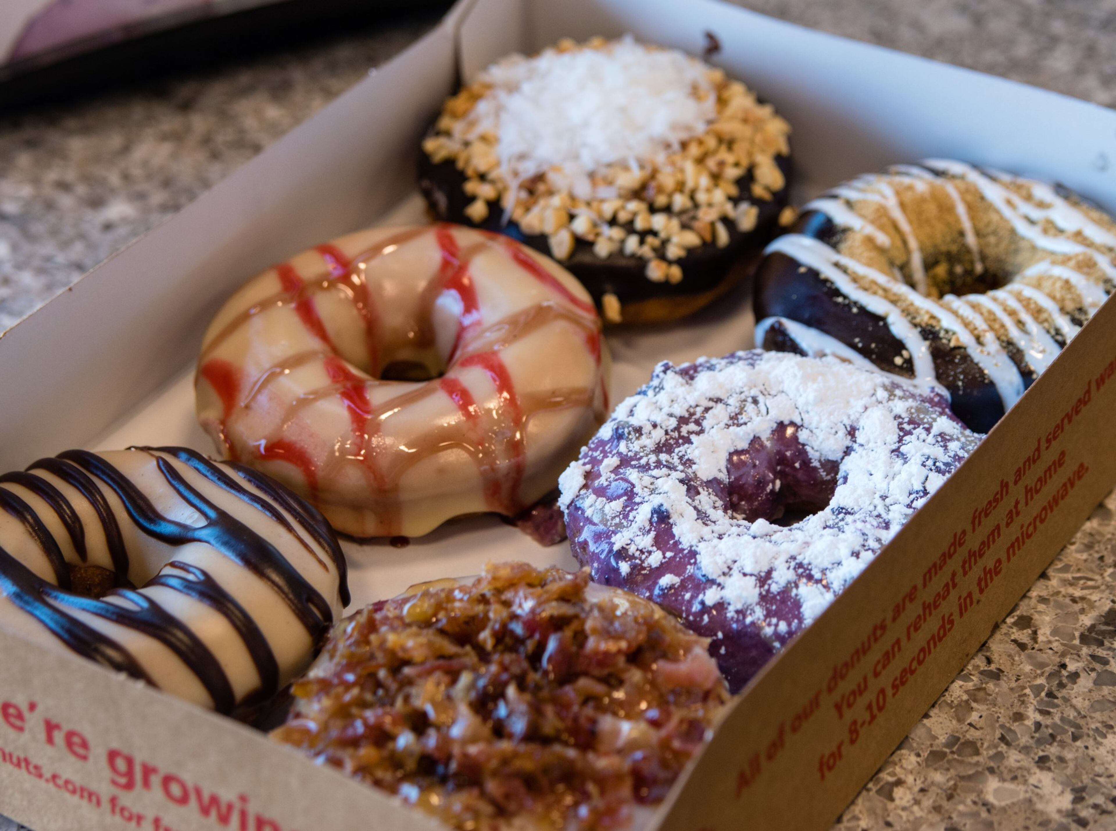 A box of six donuts at Duck Donuts (clockwise from top): Coconut Island Bliss, S’Mores, Blueberry Pancake, Bacon in the Sun, Peanut Butter Paradise and Sunrise with blackberry drizzle. (Henri Hollis/AJC)