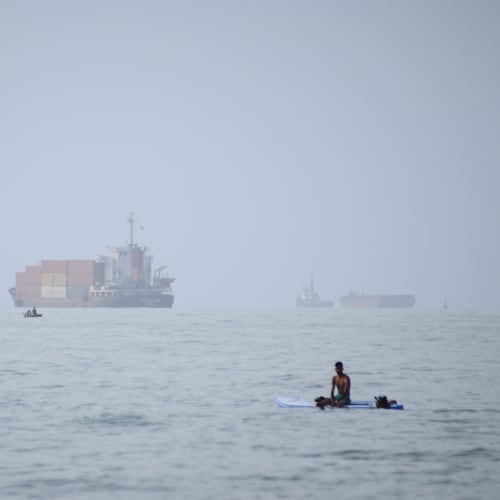 FILE - A man sits on a surfboard as a container ship and other commercial vessels appear anchored in the Strait of Hormuz off Bandar Abbas, Iran, Monday, July 27, 2026. (Razieh Poudat/ISNA via AP, File)
