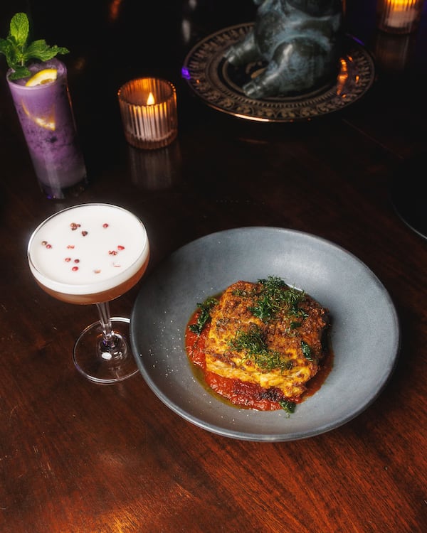 A plate of pastitsio, or Greek lasagna, with a cocktail called the Repeat Offender to the left. The purple cocktail is called the Selina Kyle. (Braxton Moore, courtesy of Buddy Buddy)
