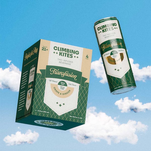 A single can of Climbing Kites Transfusion and a cardboard box labeled with the same are shown "flying" in a blue sky with white fluffy clouds.