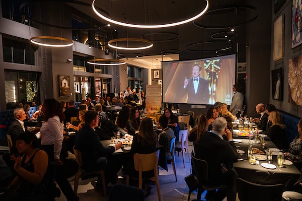 A crowd of nicely dressed people sit at dining tables while watching the Oscar's on a projector.