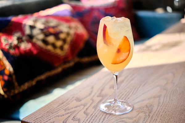 A white wine glass is full of ice and a light-colored cocktail. Floating in the glass are several peach slices.