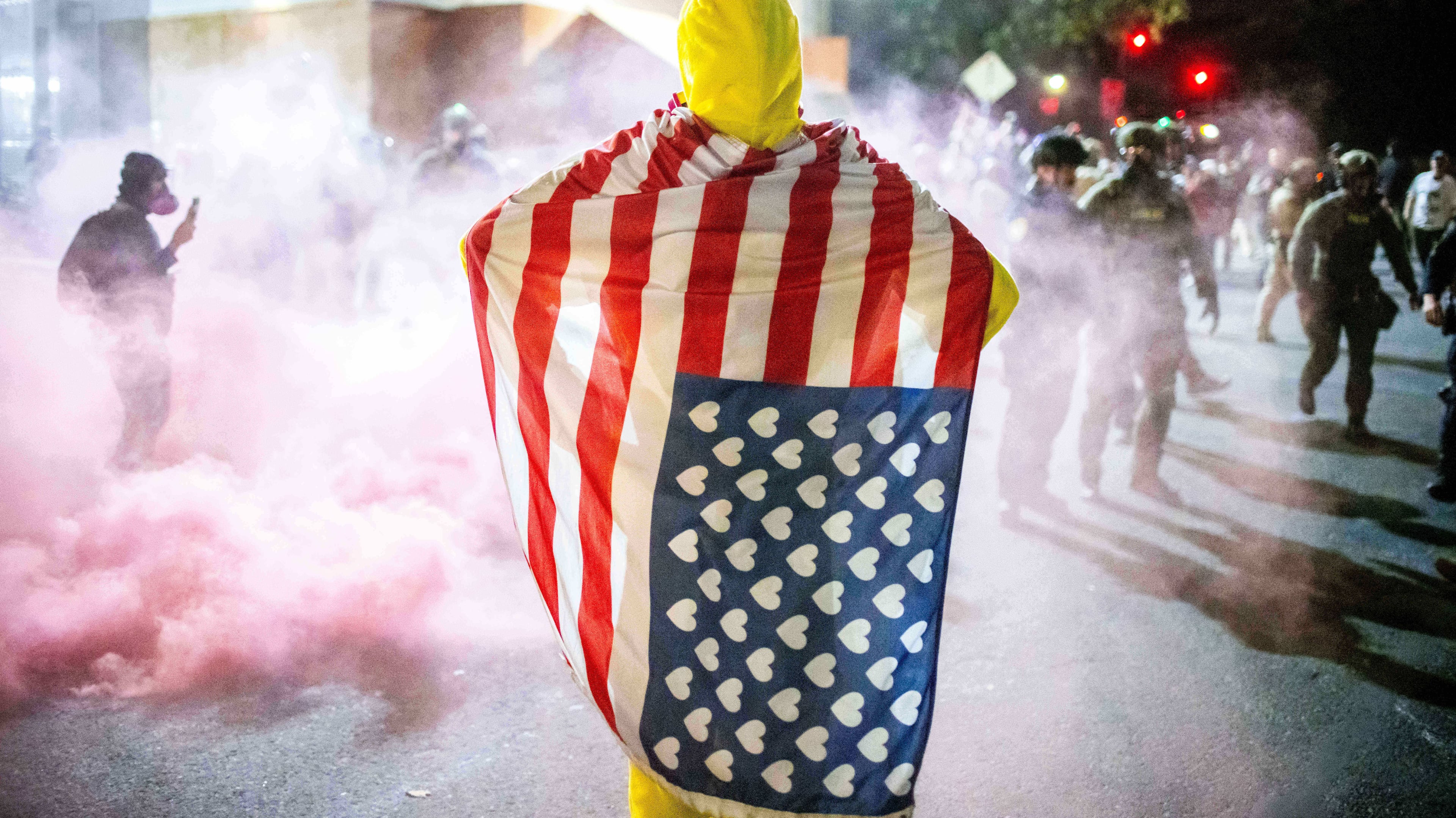 A protester, draped in an altered American flag, watches as law enforcement officers try to disperse protesters near a U.S. Immigration and Customs Enforcement facility in Portland, Ore. on Sunday, Oct. 5, 2025. (AP Photo/Ethan Swope)
