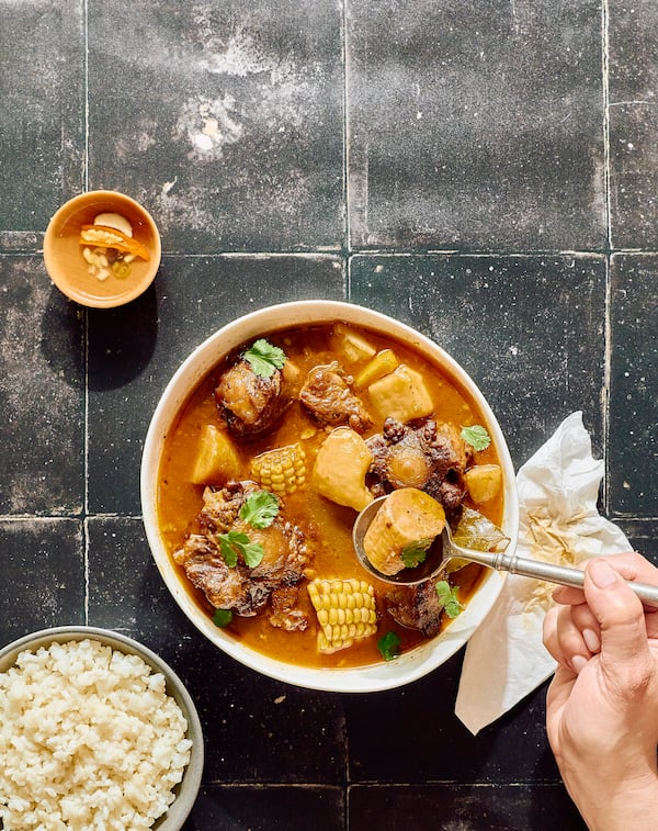 A hand holding a spoon lifts a piece of cooked corn on the cob from a bowl full of oxtail sancocho. A bowl of white rice and a small bowl of garnish are also in the image.