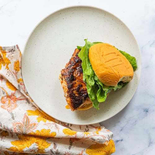 A plate with a single blackened fish sandwich and crisp lettuce on a potato roll. A colorful, floral napkin is tucked between the plate and the marble countertop.