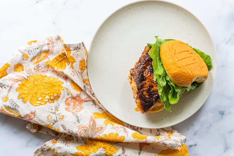 A plate with a single blackened fish sandwich and crisp lettuce on a potato roll. A colorful, floral napkin is tucked between the plate and the marble countertop.