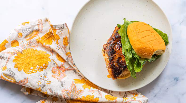A plate with a single blackened fish sandwich and crisp lettuce on a potato roll. A colorful, floral napkin is tucked between the plate and the marble countertop.