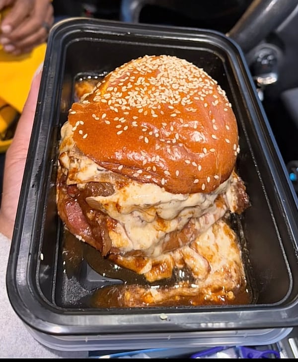 A hand holds a carryout contain containing a juicy, overflowing burger on a grilled sesame seed bun. The burger has many grilled onions and a gooey white sauce - possibly made of cheese.