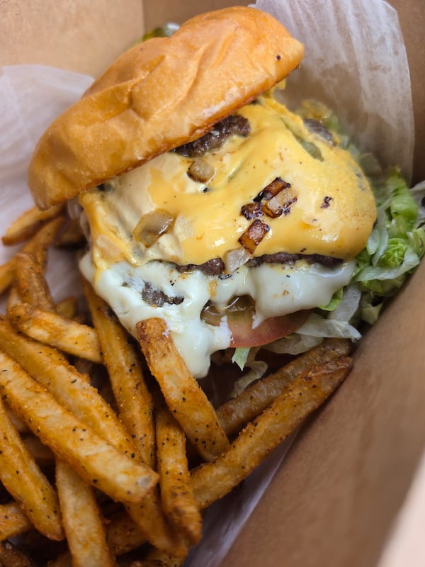 A juicy burger dripping with cheese sauce and a fried egg, nestled next to a generous amout of crispy-looking french fries. The burger is on a grilled plain bun, and is garnished with what looks like diced sauted onion, lettuce and tomato slices.