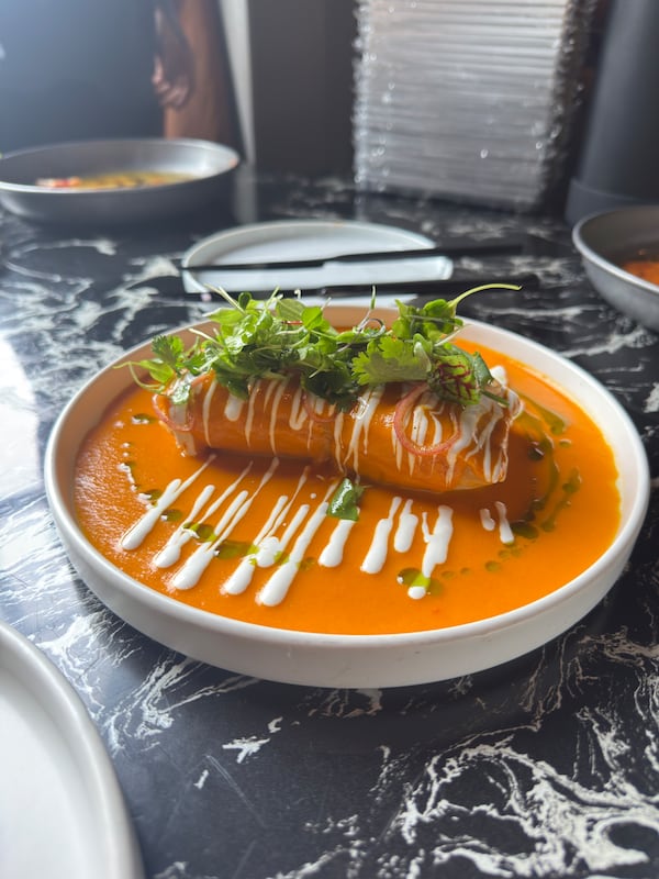 On a marble table, there is a plate full of an orange chupe sauce, drizzles of cheese and a chicken tamale with micro greens on top.