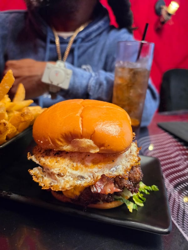 A large smash burger with a plain bun, fried egg and a little lettuce peeking out is accompanied by french fries. They are on a plate on a restaurant table. A man wearing a nice sweatshirt and stylish jewelry is in the background of the photo, resting his arms on the table. A coke is next to his elbow.