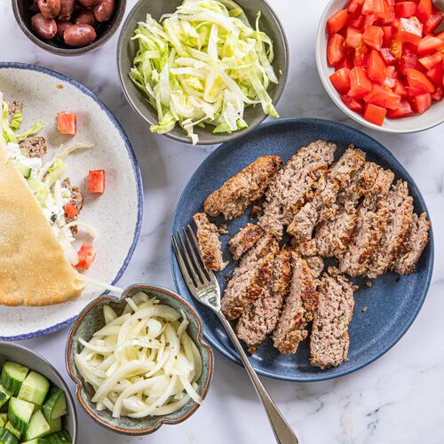 A collection of dishes is filled with ingredients that can be added to lamb gyros. There are bowls of chopped cucumbers, sliced onions, whole olives, shredded lettuce and diced tomatoes, as well as a plate of sliced gyro meat. A second plate shows an assembled gyro, with meat, lettuce and cucumber tucked into half of a toasted pita bread.