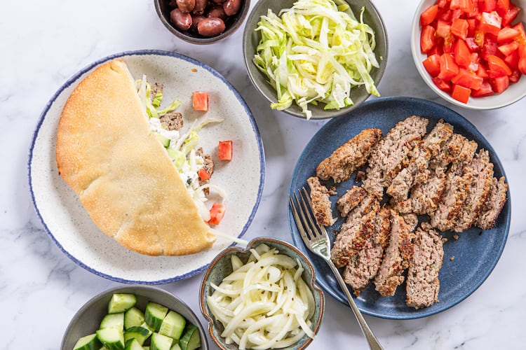 A collection of dishes is filled with ingredients that can be added to lamb gyros. There are bowls of chopped cucumbers, sliced onions, whole olives, shredded lettuce and diced tomatoes, as well as a plate of sliced gyro meat. A second plate shows an assembled gyro, with meat, lettuce and cucumber tucked into half of a toasted pita bread.