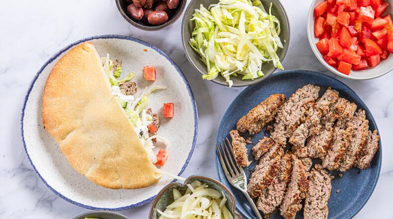 A collection of dishes is filled with ingredients that can be added to lamb gyros. There are bowls of chopped cucumbers, sliced onions, whole olives, shredded lettuce and diced tomatoes, as well as a plate of sliced gyro meat. A second plate shows an assembled gyro, with meat, lettuce and cucumber tucked into half of a toasted pita bread.
