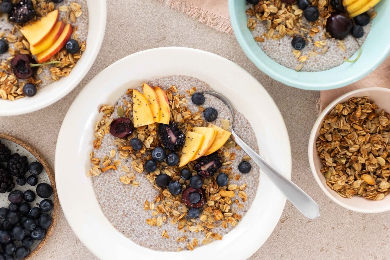 Overhead view of a chia pudding bowl topped with blueberries, peaches and granola, with more bowls of chia pudding extending out of the frame and additional bowls of granola and berries.