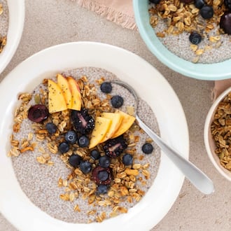 Overhead view of a chia pudding bowl topped with blueberries, peaches and granola, with more bowls of chia pudding extending out of the frame and additional bowls of granola and berries.