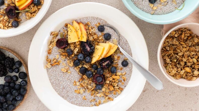 Overhead view of a chia pudding bowl topped with blueberries, peaches and granola, with more bowls of chia pudding extending out of the frame and additional bowls of granola and berries.