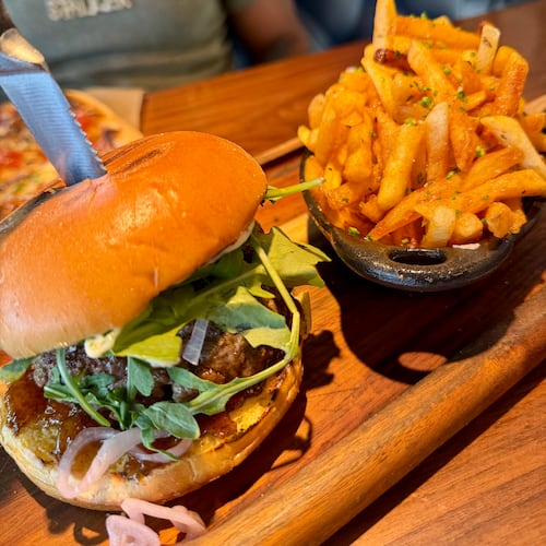 A burger and fries served on a wood plank. The thick burger has both carmelized onions and sliced raw shallots. It has arugula mixed greens and a smooth bun. A steak knife is stabbed into the center of the burger. The french fries are lightly golden and a sprinkle of fresh green herbs on top.