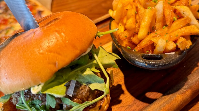 A burger and fries served on a wood plank. The thick burger has both carmelized onions and sliced raw shallots. It has arugula mixed greens and a smooth bun. A steak knife is stabbed into the center of the burger. The french fries are lightly golden and a sprinkle of fresh green herbs on top.