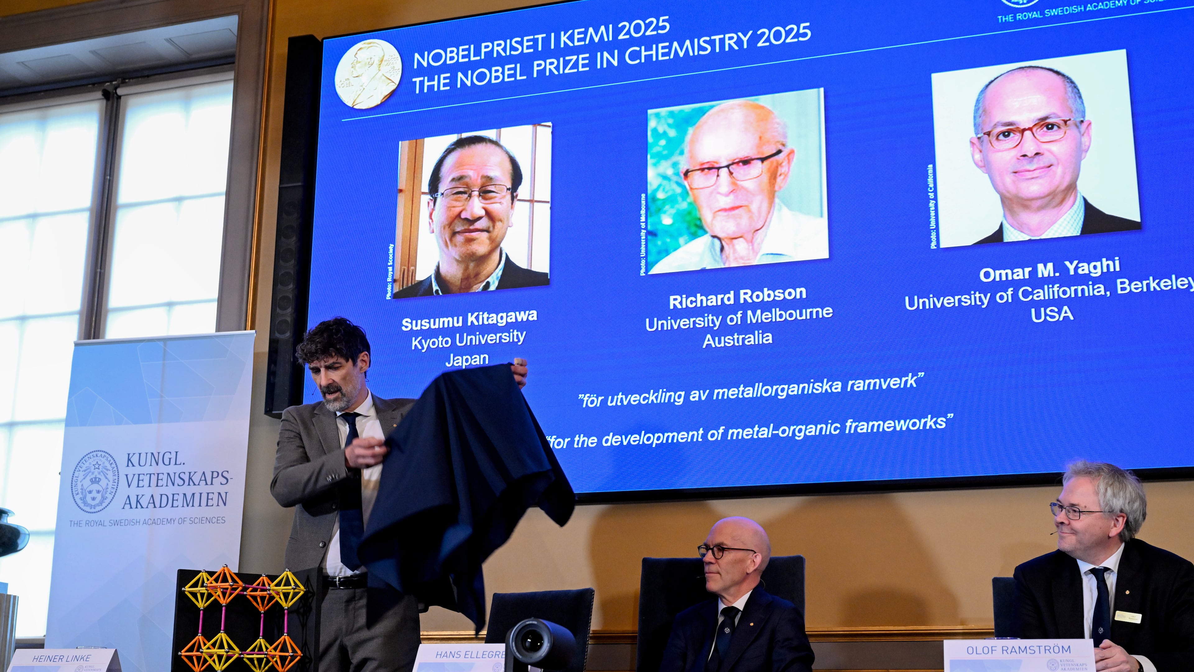 Chair of the Nobel Committee for Chemistry Heiner Linke makes a demonstration, next to Secretary General of the Swedish Academy of Sciences Hans Ellegren, and Member of the Nobel Committee for Chemistry Olof Ramstrom, right, after they announce Susumu Kitagawa, Richard Robson and Omar Yaghi, on screen behind, as the recipients the Nobel Prize in Chemistry, at the Nobel Assembly of the Karolinska Institutet, in Stockholm, Sweden, Wednesday, Oct. 8, 2025. (Fredrik Sandberg/TT News Agency via AP)