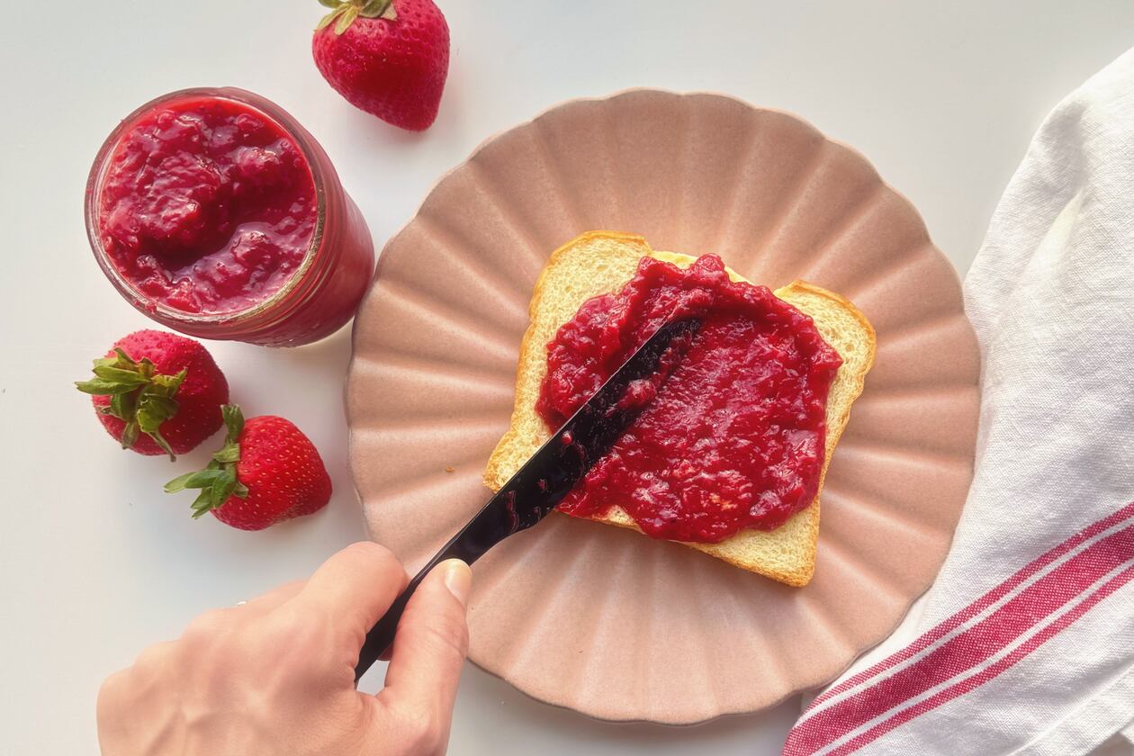 A hand holds a black butter knife and spreads strawberry quick jam on a slice of brioche bread that's set on a pink plate with an open jar of strwberry quick jam to the left opf the hand surrounded by fresh strawberries and a white cloth napkin with a red stripe to the right of the pink plate.