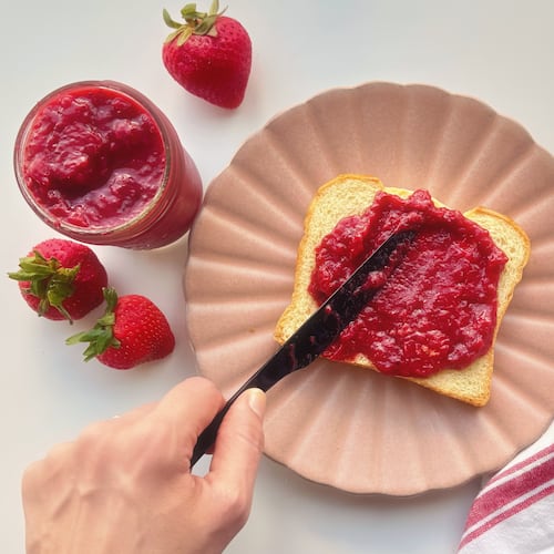 A hand holds a black butter knife and spreads strawberry quick jam on a slice of brioche bread that's set on a pink plate with an open jar of strwberry quick jam to the left opf the hand surrounded by fresh strawberries and a white cloth napkin with a red stripe to the right of the pink plate.