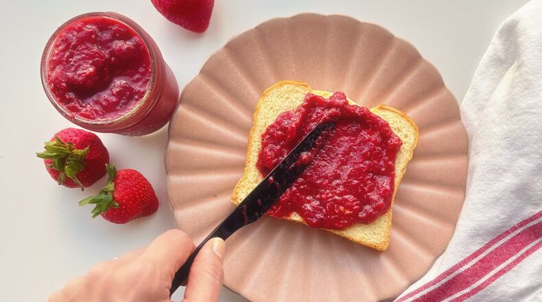 A hand holds a black butter knife and spreads strawberry quick jam on a slice of brioche bread that's set on a pink plate with an open jar of strwberry quick jam to the left opf the hand surrounded by fresh strawberries and a white cloth napkin with a red stripe to the right of the pink plate.
