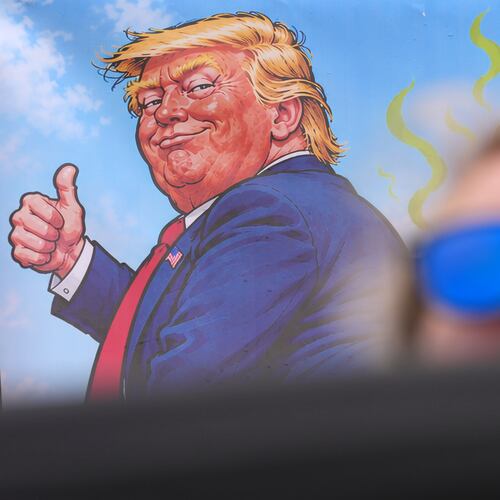A protester wears sunglasses and sits in front of a poster depicting U.S. President Donald Trump during his visit to the Irish Open at the Trump International Golf Links course in Doonbeg, Ireland, Sunday, Sept. 13, 2026.(AP Photo/Kin Cheung)