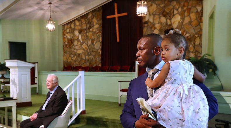 The Rev. Tony Lowden holds his daughter, Tabitha, as he stands in the sanctuary of Maranatha Baptist Church in Plains, Georgia as he and former President Jimmy Carter, seated, wait to greet visitors following a worship service in June 2019. (Curtis Compton/The Atlanta Journal-Constitution/TNS)