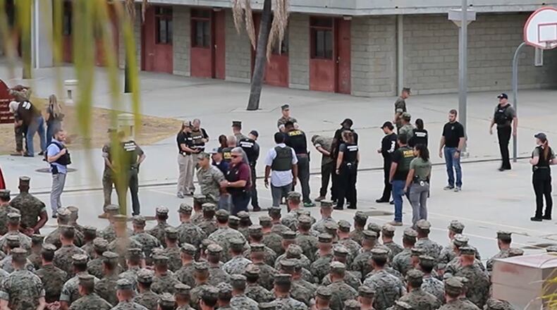 A screenshot shows military law enforcement, including NCIS, handcuff, search and walk detained Marines in front of their peers at Marine Corps Base Camp Pendleton in California.
