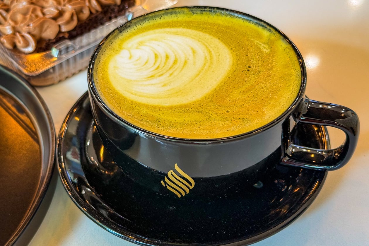 An artfully poured latte in a black mug sits on a saucer on a table in a coffee shop.