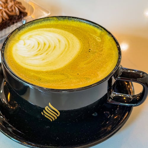 An artfully poured latte in a black mug sits on a saucer on a table in a coffee shop.