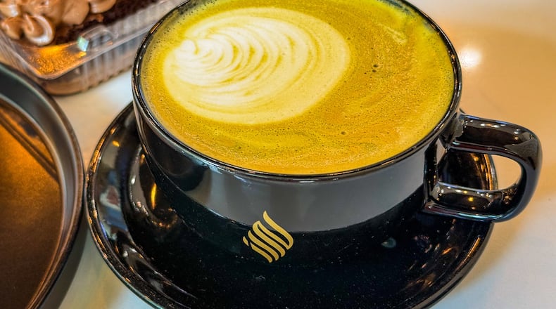 An artfully poured latte in a black mug sits on a saucer on a table in a coffee shop.
