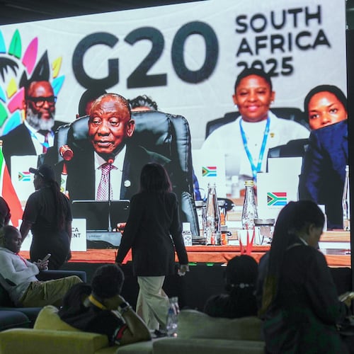 People walk by a large screen TV where South African President Cyril Ramaphosa holds a wooden gavel as he officially closes the G20 leaders' summit, in Johannesburg, South Africa, Sunday, Nov. 23, 2025. (AP Photo/Jerome Delay)