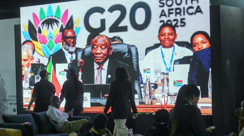 People walk by a large screen TV where South African President Cyril Ramaphosa holds a wooden gavel as he officially closes the G20 leaders' summit, in Johannesburg, South Africa, Sunday, Nov. 23, 2025. (AP Photo/Jerome Delay)