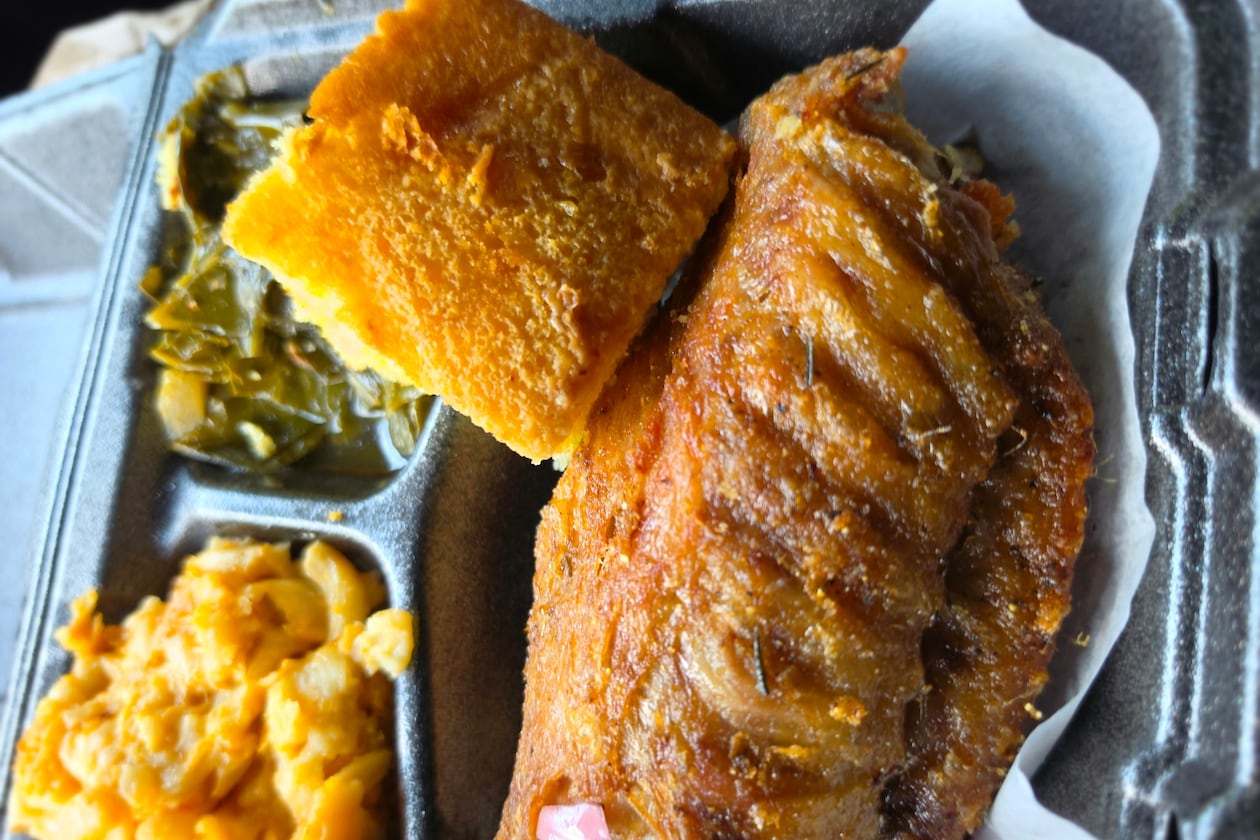 A close-up shot of a takeout container shows lemon pepper turkey wing, with sides of macaroni and cheese and collard greens tucked into the tray compartments. A square of cornbeard balances on top of the turkey wing.