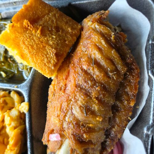 A close-up shot of a takeout container shows lemon pepper turkey wing, with sides of macaroni and cheese and collard greens tucked into the tray compartments. A square of cornbeard balances on top of the turkey wing.