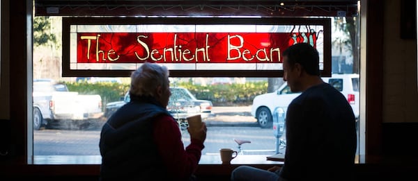 Two people are silhouetted by an exterior shop window with a stained glass sign that reds "The Sentient Bean."