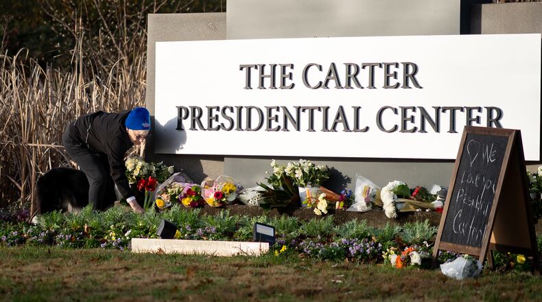 Laura Cole places amaryllis flowers on the sign at the Carter Presidential Center in Atlanta on Monday, December 30, 2024. (Ben Hendren for the Atlanta Journal-Constitution)