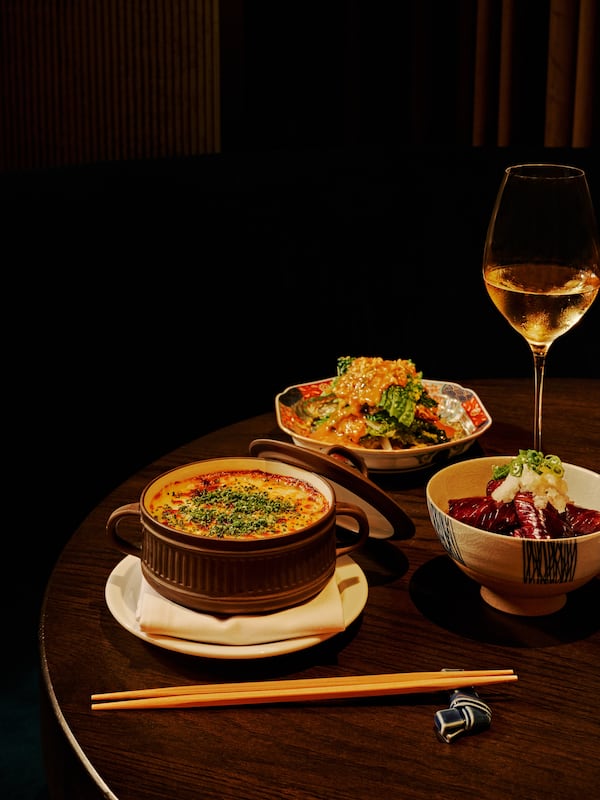 Three dishes of cooked japanese vegetables sit on a table in a dark room with chopsticks and a glass of wine.